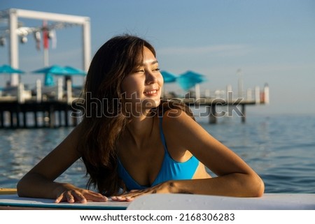 Portrait Asians beautiful girl in a blue bikini  sits on a surfboard and laughs. in the ocean against the backdrop of sunset. Woman on paddleboarding