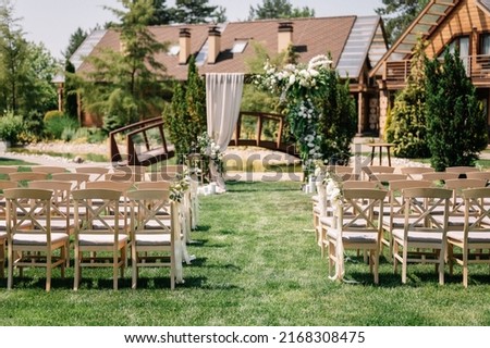 Wedding ceremony. On a green meadow in the forest there is an arch of flowers and greenery, next to there are wooden chairs for guests