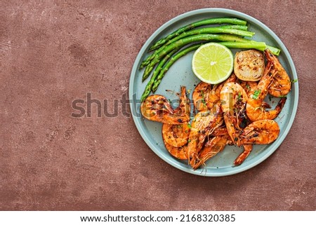 Grilled shrimp (tiger prawns) in spices with asparagus and a slice of lime (lemon) on a plate on a brown background.