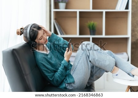 Peaceful girl in modern wireless headphones sit relax on comfortable couch listening to music, Asian young woman listening to music with smartphone and relaxing while sitting on sofa at home.