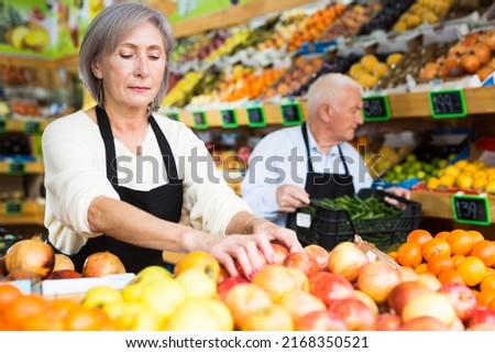 Woman merchandiser in apron putting goods on shelf in greengrocer