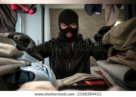 a thief in a black mask searches a wardrobe in search of money and valuables. concept of apartment theft. stealing clothes. Royalty-Free Stock Photo #2168384415