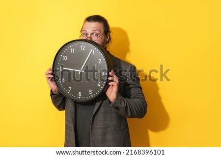 Young businessman peeking out from behind big clock on a yellow background