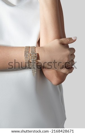 Cropped close-up shot of a woman with a gold rhinestone bracelet with a buckle. The woman in a white blouse is wearing a rhinestone bracelet on a light background. Front view. Royalty-Free Stock Photo #2168428671