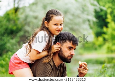 Side view of wonderful family sitting on picnic in park forest around trees bushes. Little discontented girl daughter sitting on fathers back. Middle-aged man eating food. Love, summer, togetherness.