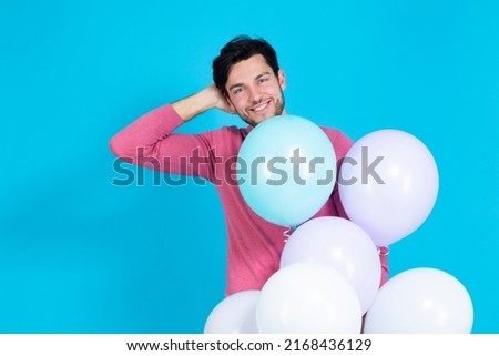 One Caucasian Guy Handsome Brunet Man With Bunch of Colorful Air Balloons in Pink Jumper Standing With Lifted Hand Behind On Blue Background. Horizontal image