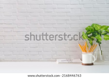Comfortable workplace with blank picture frame, coffee cup, book and stationery on white table against brick wall. Copy space for your text.