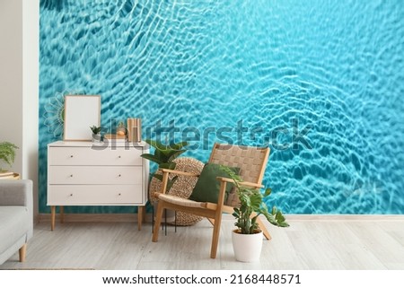 Comfortable armchair, chest of drawers and houseplants near wall with print of clear blue water