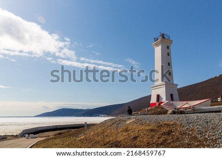 Lighthouse (leading beacon) and a boat on the shore of the sea bay. View from the shore to the ice-covered bay. Nagaev Bay, Sea of Okhotsk. Park Lighthouse in the city of Magadan. Far East of Russia. Royalty-Free Stock Photo #2168459767