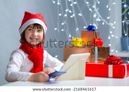 One Winsome Caucasian Child Girl In Santa Hat With Cellphone Using Laptop for Online Gift Search While Using Video Calls By Webcam Chat During Holidays. Horizontal Image