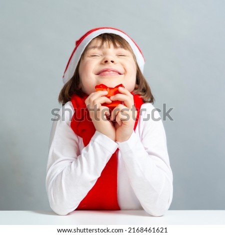 Holidays Concepts. One Dreaming Winsome Caucasian Female Kid in Santa Hat and White Shirt Holding Tiny Red Gift Box in Front of Face. Square Image