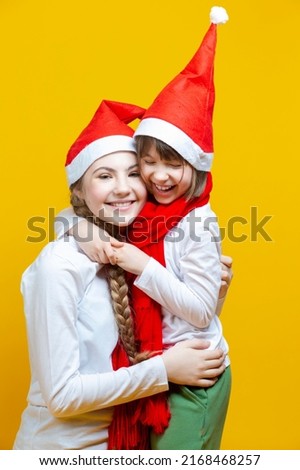 Family of Two Positive Caucasian Girls Sisters  In Festive Santa Hats Having Fun While Embracing And Toching by Cheeks On Yellow Background. Vertical image