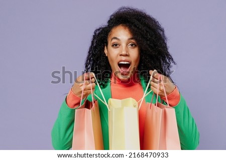 Shocked amazed excited fun young black curly woman 20s wears green shirt look camera holding package bags with purchases after shopping isolated on plain pastel light violet background studio portrait