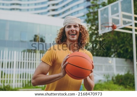 Young happy fun cool man 20s wearing yellow t-shirt bandana hold ball playing basketball on playground in free time walking rest relax in city outdoors on open air. Urban lifestyle leisure concept.