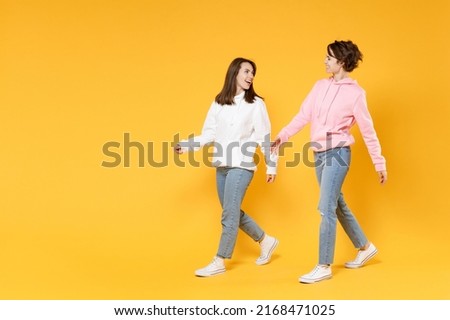 Full length side view of laughing funny two young women friends 20s wearing casual white pink hoodies walking going looking at each other isolated on bright yellow color background studio portrait