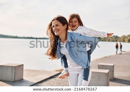 Embracing each other and smiling. Young mother with her daughter having fun outdoors near the lake at summer.