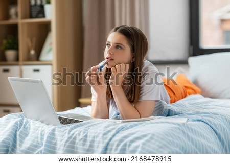 school, online education and e-learning concept - thinking or daydreaming teenage student girl with laptop computer writing to notebook lying on bed at home Royalty-Free Stock Photo #2168478915