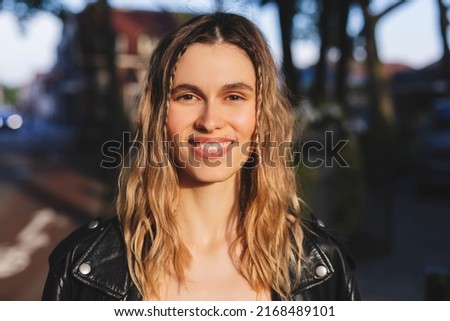 Pensive blonde woman in black leather jacket posing on blur city street background. Outdoor shot of happy hippie lady with two thin braids and wave hair. Coachella or boho freedom style. Royalty-Free Stock Photo #2168489101