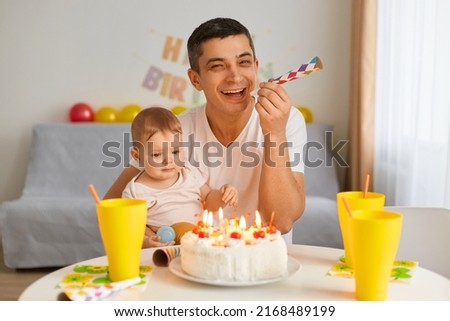 Image of young adult man with toddler baby celebrating first birthday, sitting at table, looking at birthday cake, father holding party horn, laughing, posing with his charming cute kid.