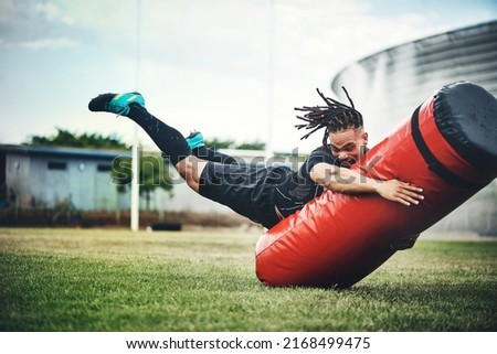 He trains like the rugby pro he is. Full length shot of a handsome young rugby player working out with a tackle bag on the playing field. Royalty-Free Stock Photo #2168499475