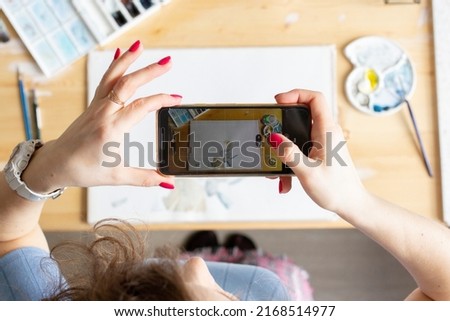 Woman taking photo of her art watercolor picture, aquarelle process, girl blogger making contet for art blog, artist workplace with watercolors, brushes and paper, mobile phone in women hands top view