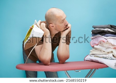 A funny bald man with emotions using electrical iron on ironing board over on blue background.