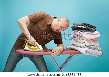 view of funny bald man, he tries to iron his shirt on himself on blue background.