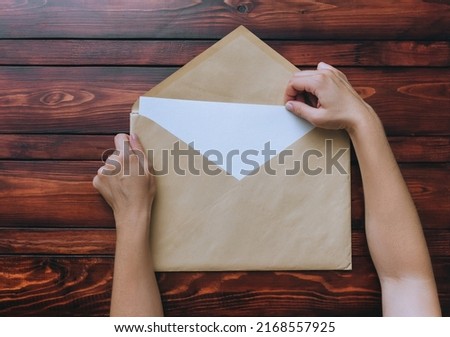 Two female hands hold a large kraft beige kraft paper envelope with a blank white sheet of paper inside lies on a brown wooden background. Blank letter concept with copy space.