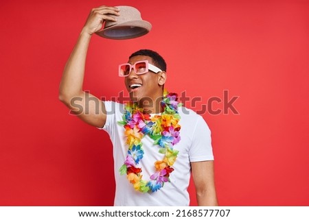 Happy young man in Hawaiian necklace carrying hat while standing against red background                      