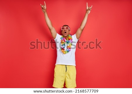 Happy man in Hawaiian necklace keeping arms raised while standing against red background        