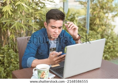 A young asian man disturbed by a controversial video on his cellphone. Distracted with social media during a remote work session at an outdoor cafe. Royalty-Free Stock Photo #2168586555