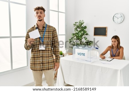 Young handsome man voting putting envelop in ballot box thinking attitude and sober expression looking self confident 