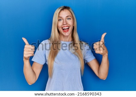 Beautiful blonde woman wearing casual t shirt over blue background success sign doing positive gesture with hand, thumbs up smiling and happy. cheerful expression and winner gesture. 