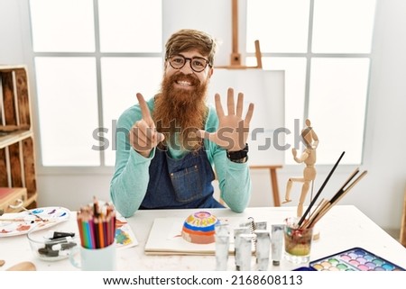 Redhead man with long beard painting clay bowl at art studio showing and pointing up with fingers number six while smiling confident and happy. 