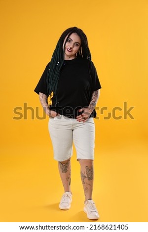 Beautiful young woman with tattoos on body, nose piercing and dreadlocks against yellow background