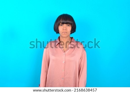 Joyful young brunette woman wearing pink silk shirt over blue background looking to the camera, thinking about something. Both arms down, neutral facial expression.