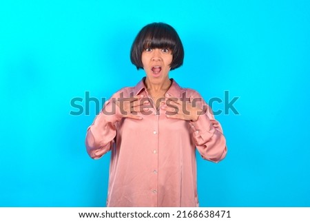 young brunette woman wearing pink silk shirt over blue background keeps hands on chest feeling shocked and scared, mouth widely opened, stares at camera saying: Who, me?
