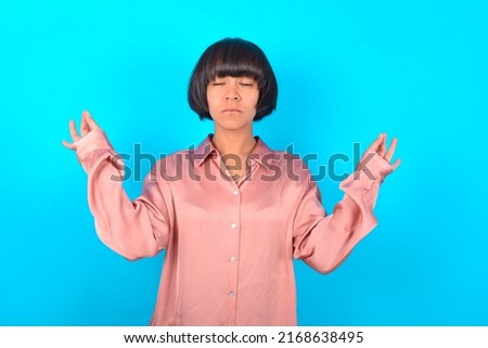 young brunette woman wearing pink silk shirt over blue background doing yoga, keeping eyes closed, holding fingers in mudra gesture. Meditation, religion and spiritual practices.