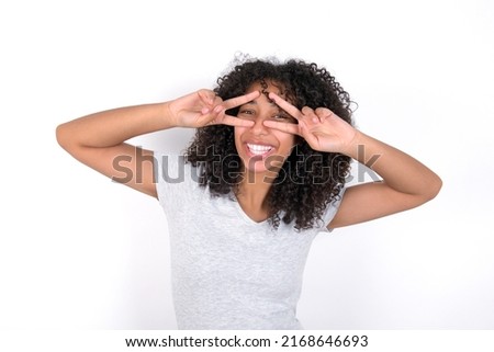 Cheerful positive Young beautiful girl with afro hairstyle wearing grey t-shirt over white background shows v-sign near eyes open mouth