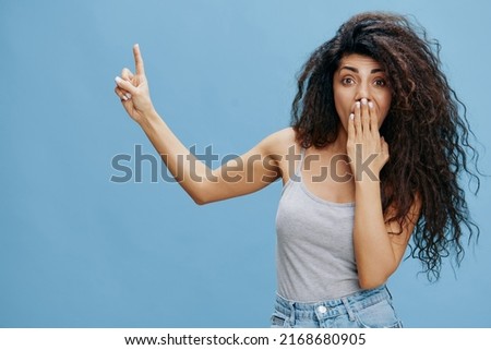 COOL JEANS OFFER. Amazed beautiful Latin female with afro close mouth, point finger up, look at camera. Fashion. Studio shoot over blue sky background. Copy space, free place for design ad