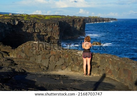 Blonde girl taking some pictures of the cliffs of lava rock plummeting into the Pacific Ocean located on the southern side of the Hawaiian Volcanoes National Park on the Big Island of Hawai'i