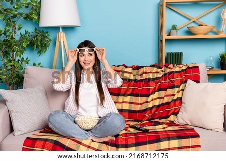 Portrait of attractive trendy cheerful girl sitting on divan watching film wear spectacles isolated on blue color background indoors