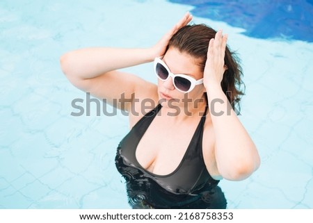 Stylish young woman plus size body positive in black swimsuit and white sunglasses swimming in the pool, summer vacations