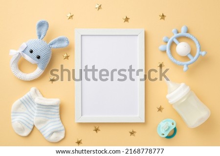 Baby accessories concept. Top view photo of photo frame knitted bunny rattle toy blue teether milk bottle tiny socks baby's dummy and gold stars on isolated pastel beige background with blank space