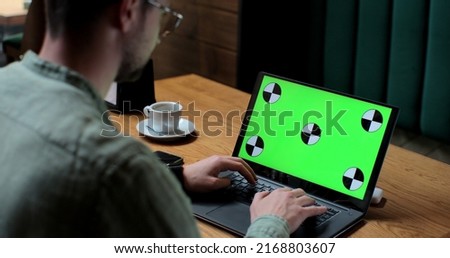 Young man in glases working on laptop with green screen in coffee shop.