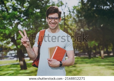 Young student man in blue t-shirt eyeglasses backpack he hold books show v-sign walk rest relax in spring green city park go down alley sunshine lawn outdoors on nature. Education high school concept