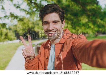 Close up young smiling man in orange jacket walk do selfie shot pov mobile phone show v-sign rest relax in spring green city park go down alley sunshine lawn outdoor on nature Urban leisure concept.