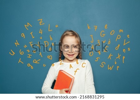 Happy smiling kid girl with book having fun against blue background with letters