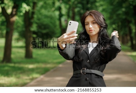 Young woman in a black suit makes selfie while working outdoors, walking in the park, communicating by video link