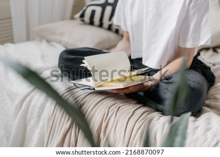 Cropped view of calm woman reading book in bright bedroom. Female sitting on bed, turning pages in her notepad with empty pages. Inspiration concept. Morning in room with bohemian chic style Royalty-Free Stock Photo #2168870097